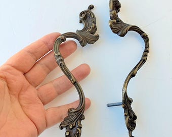 May include: Two antique bronze-colored cabinet handles with ornate, curved designs. Each handle features a scrolling leaf pattern and a decorative finial. The handles are shown against a white background.