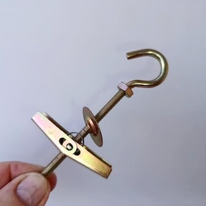 Puede incluir: Un perno de palanca de metal en tono dorado que presenta una varilla roscada con un gancho en un extremo, una arandela circular y un mecanismo de ala de palanca con resorte en el otro extremo. El herraje tiene un acabado metálico brillante.