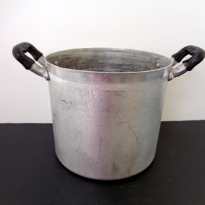 Puede incluir: Una olla de cocina grande y cilíndrica de aluminio con dos asas negras. La olla tiene un acabado plateado y una banda decorativa cerca de la parte superior. La olla está diseñada para cocinar y preparar alimentos.