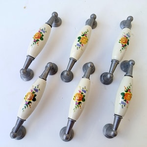 May include: Six vintage cabinet handles. Each has a cream ceramic centre with a floral design in yellow, orange, and purple. The handles have a silver metal base and mounting hardware. Ideal for kitchen or furniture.