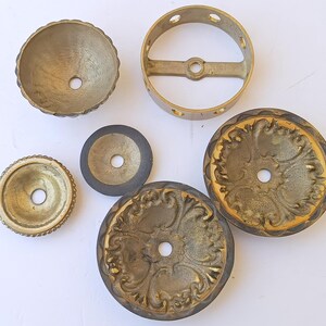 May include: A collection of six brass-toned metal hardware components, including two ornate circular plates with embossed floral patterns, a small bowl-shaped piece, a flat washer, a plain disc, and a circular ring with four side holes.