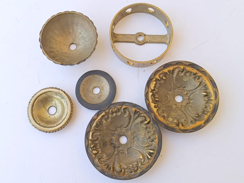 May include: A collection of six brass-toned metal hardware components, including two ornate circular plates with embossed floral patterns, a small bowl-shaped piece, a flat washer, a plain disc, and a circular ring with four side holes.