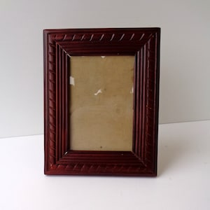 Könnte beinhalten: Ein rechteckiger, dunkelroter Holzbilderrahmen mit einem dekorativen, seilartigen Muster an den Rändern. Der Rahmen hat ein schlichtes, klassisches Design und ist leer, mit einer hellbraunen Rückseite.