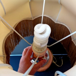 Op de afbeelding: Een witte lampenkap met een bruine franje. Een witte energiebesparende gloeilamp is in de lampvoet geschroefd.