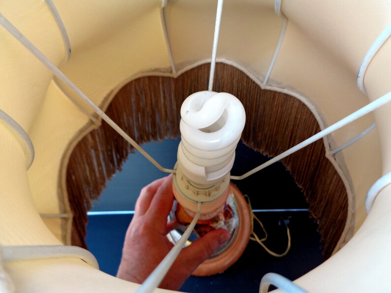 Op de afbeelding: Een witte lampenkap met een bruine franje. Een witte energiebesparende gloeilamp is in de lampvoet geschroefd.