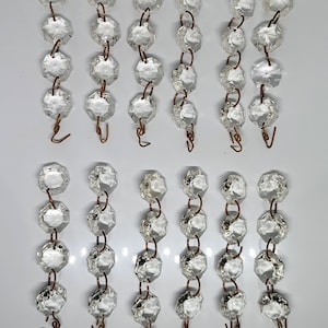 Puede incluir: Cadenas de cristal transparente y facetado para lámparas de araña, con eslabones y ganchos de alambre color cobre. Los cristales son octogonales y reflejan la luz. Las cadenas están dispuestas verticalmente sobre un fondo blanco, sugiriendo un elemento de iluminación o decoración.
