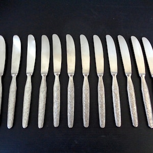 May include: A set of twelve silver butter knives arranged on a dark surface. Each knife features a curved blade and a handle with an ornate floral pattern. The knives are approximately 7 inches long.