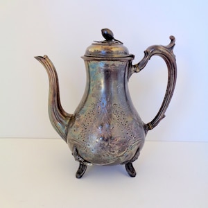 May include: Antique silver-plated coffee pot with ornate floral and scrollwork detailing. The pot features a curved spout, a decorative handle, and rests on three small feet. The lid is topped with a small, stylized finial.