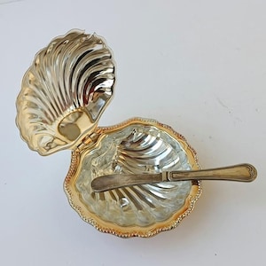 May include: A silver-toned metal clam shell-shaped dish with a hinged lid and a decorative beaded edge. The interior features a polished, reflective finish with embossed ridges. A matching silver-toned metal spreader knife rests inside the dish.