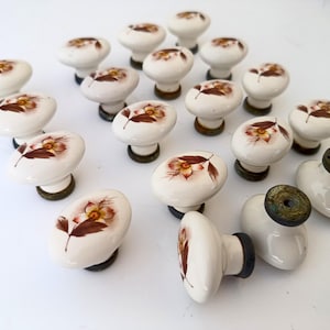 Puede incluir: Un conjunto de pomos de cerámica blanca para armarios con un diseño floral en tonos marrones y amarillos. Los pomos tienen forma redondeada y están montados sobre una base oscura. Ideal para la restauración de muebles.