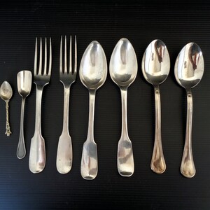 May include: A set of seven silver utensils, including a small spoon, a larger spoon, two forks, and three soup spoons. The utensils are arranged in a row on a black surface.