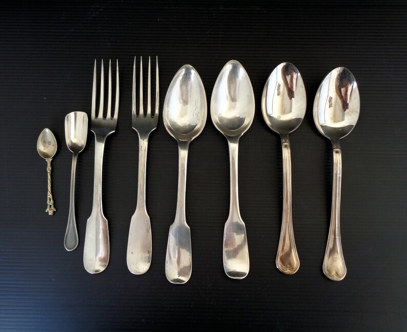 May include: A set of seven silver utensils, including a small spoon, a larger spoon, two forks, and three soup spoons. The utensils are arranged in a row on a black surface.