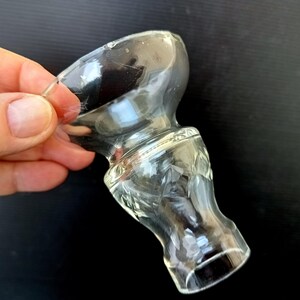 May include: A clear glass bottle stopper with a bulbous top and a decorative, etched design on the lower portion. The stopper is held against a dark background, showcasing its transparency and intricate details.