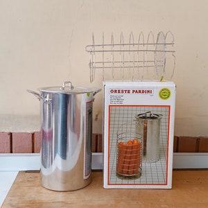 May include: A stainless steel pot with a lid, a wire rack, and a box labelled "ORESTE PARDINI" with a picture of a hot dog steamer. The box also has text in multiple languages. The items are on a wooden surface.