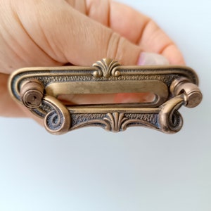 Peut inclure: Une poignée de tiroir en laiton antique avec un design orné. La poignée rectangulaire présente une ouverture centrale, des volutes décoratives et un élément floral stylisé. La quincaillerie a une esthétique vintage et une finition métallique chaude.