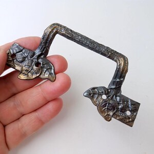 May include: A bronze-toned metal drawer pull featuring a textured branch-like handle and two mounting plates shaped like clusters of leaves. Each leaf-shaped plate contains two circular screw holes for attachment to a cabinet or furniture piece.