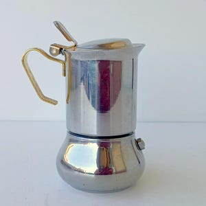 Könnte beinhalten: Eine polierte silberfarbene Moka-Kanne aus Metall mit einem goldfarbenen Griff und einem Klappdeckel mit kleinem Hebel. Der zylindrische Körper hat eine abgerundete Basis und ein kleines Überdruckventil an der Seite, das das Licht reflektiert.