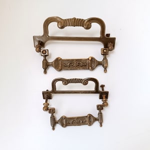 Może przedstawiać: Dwa antyczne mosiężne uchwyty do szuflad z ozdobnymi zawijasami i dekoracyjnymi panelami środkowymi. Uchwyty mają ciepłą, postarzaną patynę i zawierają elementy montażowe. Większy uchwyt ma około 15 cm szerokości, a mniejszy około 10 cm.