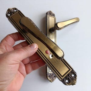 Puede incluir: Juego de manijas de puerta de latón antiguo. El juego incluye una placa rectangular larga con un ojo de cerradura y una manija adjunta. La manija tiene un diseño ornamentado. El juego se sostiene en una mano.