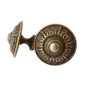 May include: An antique bronze cabinet knob with an ornate, circular design. The knob features a central, raised circle surrounded by swirling patterns. The stem and smaller, matching circular end are also visible.