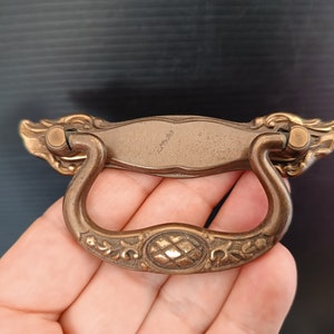 Puede incluir: Tirador de cajón de latón antiguo con detalles florales y de volutas ornamentados. El tirador es un diseño curvo, de estilo colgante, con un centro ovalado decorativo. El tirador se encuentra sobre un fondo oscuro.