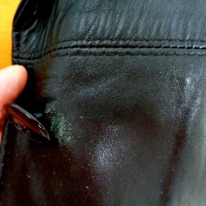 May include: Close-up of a black leather jacket with a button and stitching detail. The leather has a textured surface.