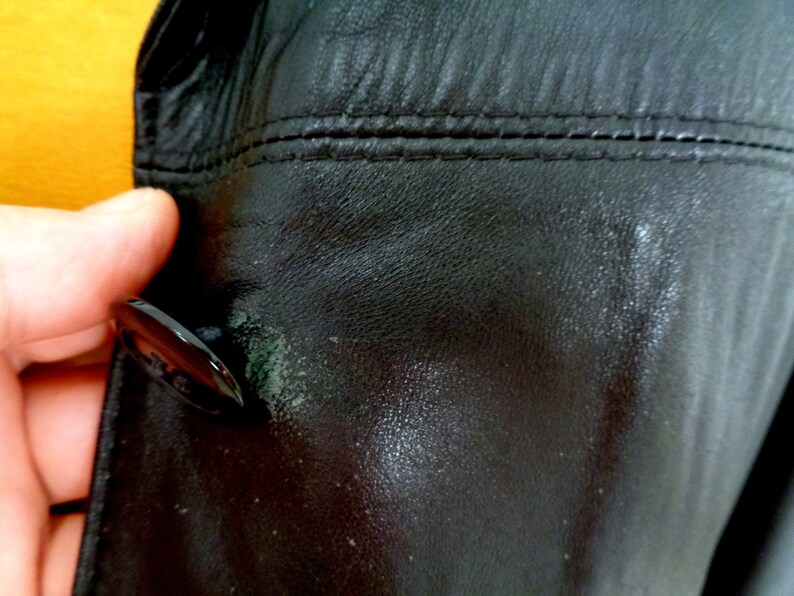 May include: Close-up of a black leather jacket with a button and stitching detail. The leather has a textured surface.
