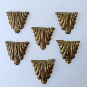 May include: Six antique brass-toned decorative furniture corner pieces. Each piece has a fan-like design with curved lines and three small holes. The pieces are arranged on a white surface.