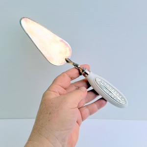 May include: A silver cake server with a white ceramic handle. The handle has a silver design with a floral pattern.