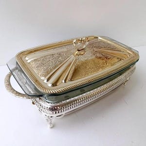 May include: A rectangular, silver-toned serving dish with a glass insert and a decorative lid. The lid features an ornate, gold-colored design with a handle. The dish sits on a silver-toned, footed base, suitable for serving food.