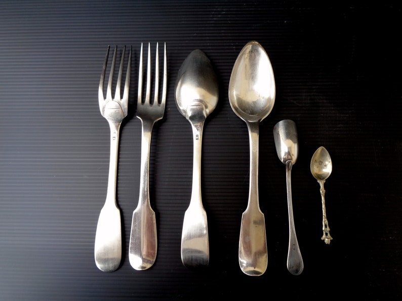 May include: A set of six silver-plated utensils, including two forks, two spoons, and two teaspoons. The utensils are arranged in a row on a black surface.