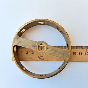 May include: A circular brass component featuring a central hub with a hole and a series of small holes along the outer rim. A wooden ruler placed behind the object indicates a diameter of approximately 3.5 inches for the brass ring structure.