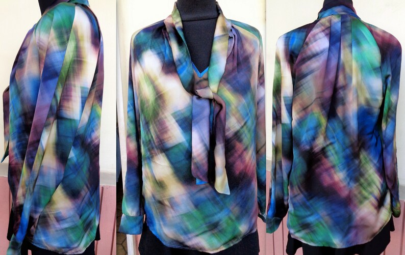 May include: A long-sleeved blouse with a colorful abstract pattern in shades of blue, green, purple, and white. The blouse features a tie-neck design and is made of a lightweight, flowing fabric. The garment is displayed on a mannequin.
