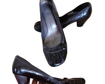 Puede incluir: Zapatos de salón negros de charol con tacón bajo y grueso. Los zapatos tienen una puntera redondeada y un detalle decorativo en la pala. El interior del zapato es de color claro. Los zapatos se muestran sobre un fondo blanco.