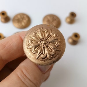 May include: Close-up of a bronze-colored cabinet knob with an ornate floral design. Several other knobs and hardware pieces are visible in the background. The knob is round and has a detailed, raised pattern.
