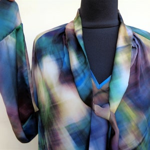 May include: A flowing, long-sleeved blouse in shades of blue, green, and purple. The fabric has a watercolor-like pattern. The blouse features a draped neckline and a contrasting blue underlayer. The sleeves are long and loose.