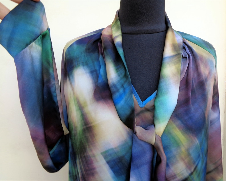 May include: A flowing, long-sleeved blouse in shades of blue, green, and purple. The fabric has a watercolor-like pattern. The blouse features a draped neckline and a contrasting blue underlayer. The sleeves are long and loose.