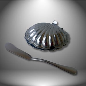 Könnte beinhalten: Eine silberfarbene Butterdose in Form einer Jakobsmuschel mit passendem Buttermesser. Die Dose hat ein geriffeltes Design und einen kleinen Griff. Das Messer hat eine abgerundete Klinge und einen gebogenen Griff. Das Set wird auf einer grauen Oberfläche präsentiert.