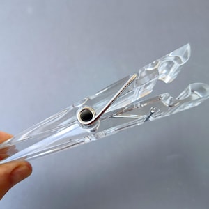 May include: A clear, transparent plastic clothespin featuring a silver-toned metal spring mechanism at its center. The clip has two long, tapered arms with grooved ends designed for gripping, showcasing a sleek, functional design.