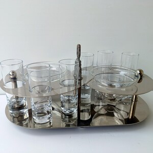 May include: A silver-toned serving tray with two tiers, holding clear glass shot glasses, small bowls, and a larger bowl. A decorative handle is centered. The tray is designed for serving appetizers or drinks.