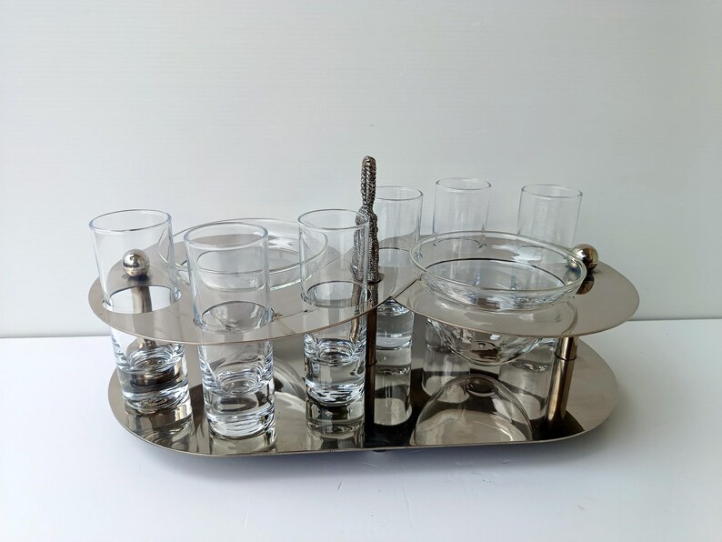 May include: A silver-toned serving tray with two tiers, holding clear glass shot glasses, small bowls, and a larger bowl. A decorative handle is centered. The tray is designed for serving appetizers or drinks.