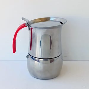 Könnte beinhalten: Ein Espressokocher aus Edelstahl mit einer polierten, reflektierenden Oberfläche. Die Kanne verfügt über einen abgerundeten Boden, einen zylindrischen oberen Behälter, einen Klappdeckel und einen geschwungenen, leuchtend roten Griff an der Seite.