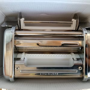 May include: A stainless steel pasta maker with white plastic components, inside a white cardboard box. The pasta machine has rollers and a handle. The text "MADE IN ITALY" is visible on the machine.