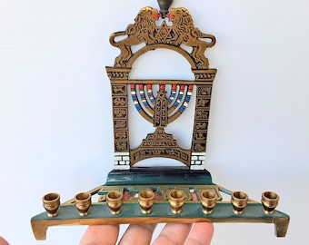 May include: A decorative menorah with a gold finish, featuring a central design with a Star of David and two lions. The menorah has eight candle holders and is painted with red, blue, and white accents. The base is green.