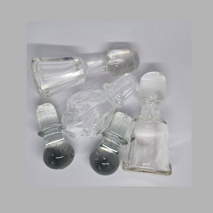 Pode incluir: Uma coleção de rolhas de garrafa de vidro transparente em várias formas e tamanhos. Algumas rolhas têm uma base bulbosa, enquanto outras têm um design mais cilíndrico. O vidro é transparente, permitindo a passagem da luz.