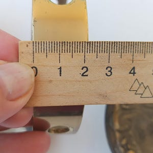 May include: A polished gold-tone metal bracket with a curved profile and circular mounting holes. A wooden ruler is positioned against the bracket, showing a width measurement of approximately 2.25 inches across the metal surface.