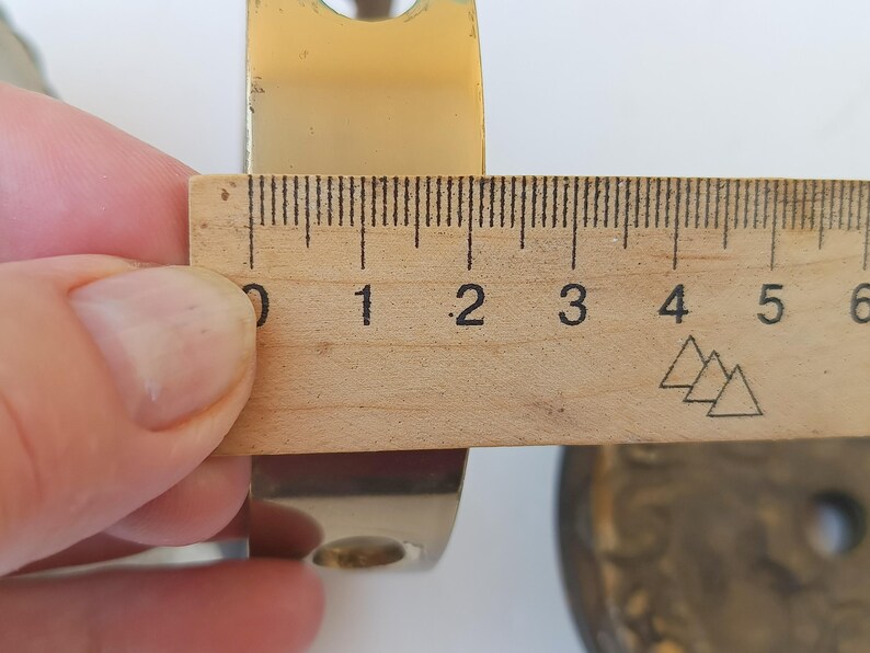 May include: A polished gold-tone metal bracket with a curved profile and circular mounting holes. A wooden ruler is positioned against the bracket, showing a width measurement of approximately 2.25 inches across the metal surface.