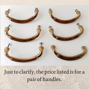 Può includere: Sei maniglie per cassetti vintage in ottone con un design curvo e un intarsio rettangolare in legno. Ogni maniglia ha una vite per una facile installazione. Le maniglie sono disposte su una superficie bianca.