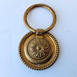 Pode incluir: Puxador de gaveta em tom de latão antigo com uma alça circular e uma placa redonda decorativa. A placa apresenta um desenho floral no centro e uma borda com contas. Um pequeno orifício é visível na parte inferior da placa.