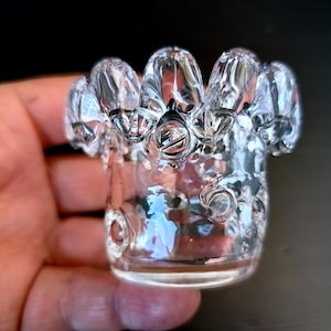Op de afbeelding: Een heldere glazen votiefkaarsenhouder met een uniek, gestructureerd ontwerp. De bovenrand heeft een reeks afgeronde, bloembladachtige vormen. Het glas heeft een bubbeltextuur, wat visuele interesse toevoegt. De houder is ontworpen om een kleine kaars te bevatten.